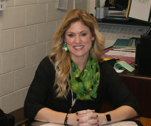 Danielle, wearing a black sweater and green scarf, sitting at her desk